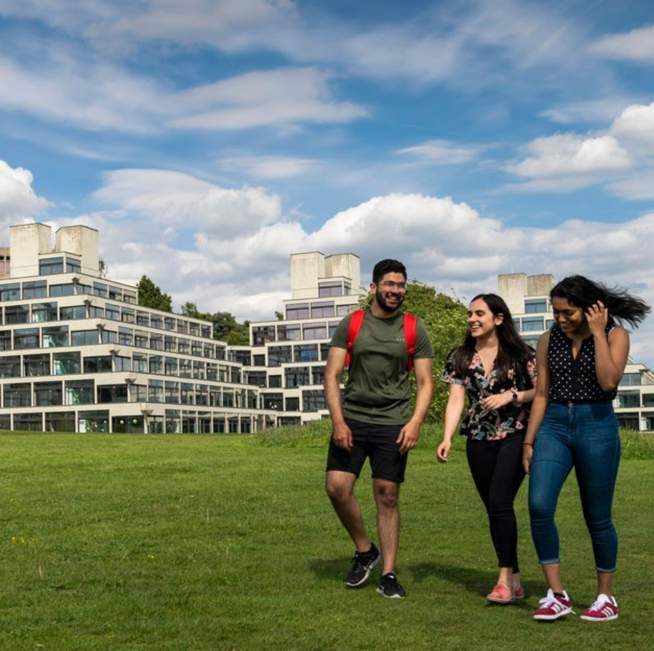 A photo of three students walking on the UEA Campus, accommodation in the background